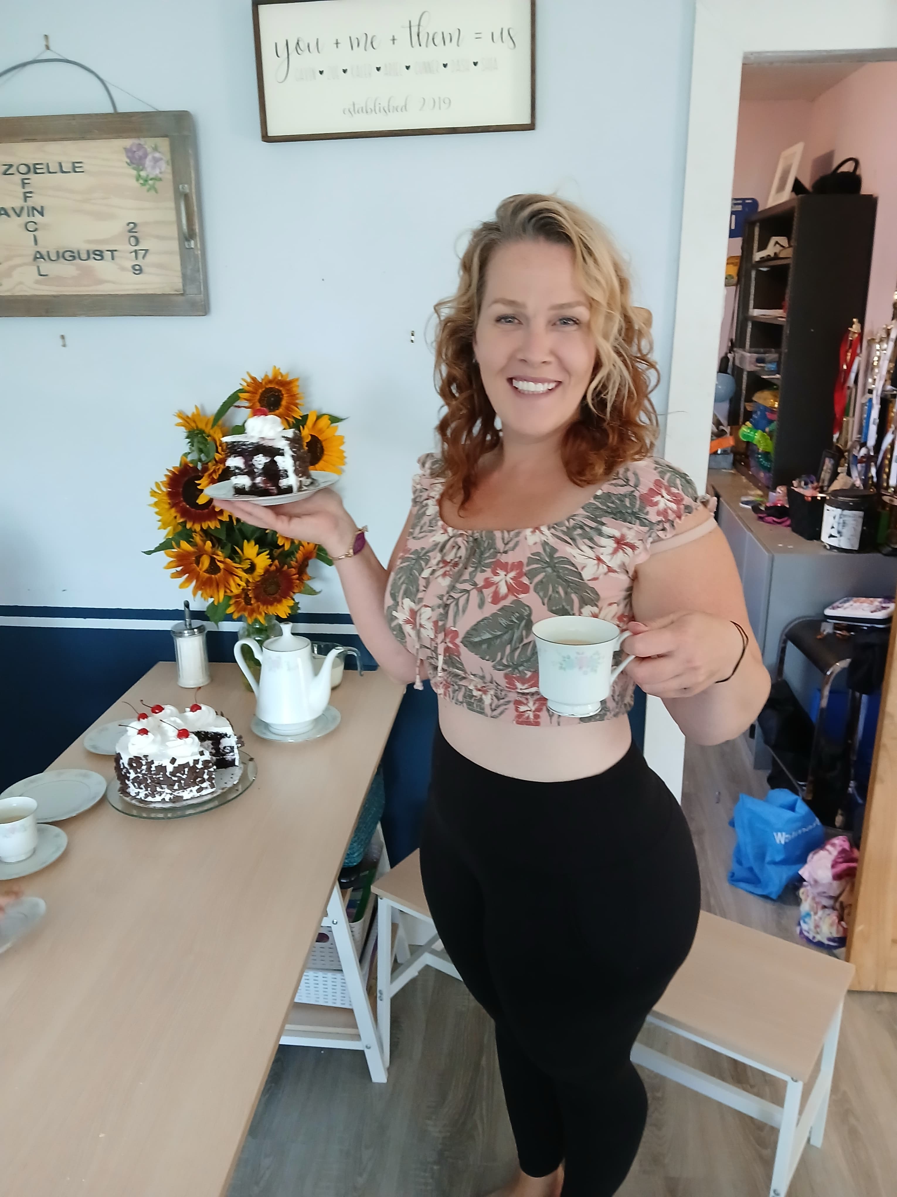 Smiling woman in floral top holding teacup and cake slice next to sunflowers and teapot.