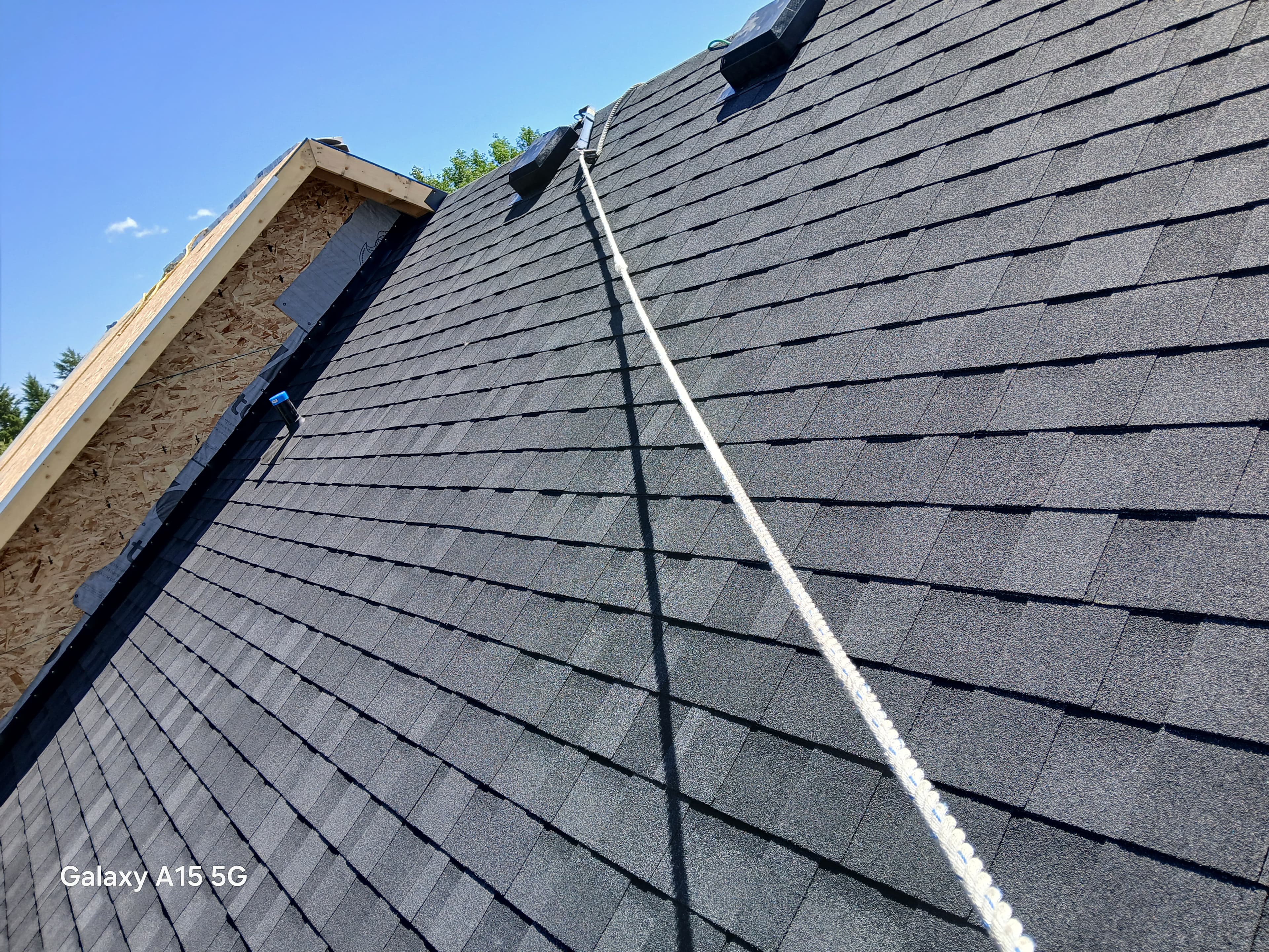 Steep dark grey shingle roof with a white safety rope and unfinished plywood section.