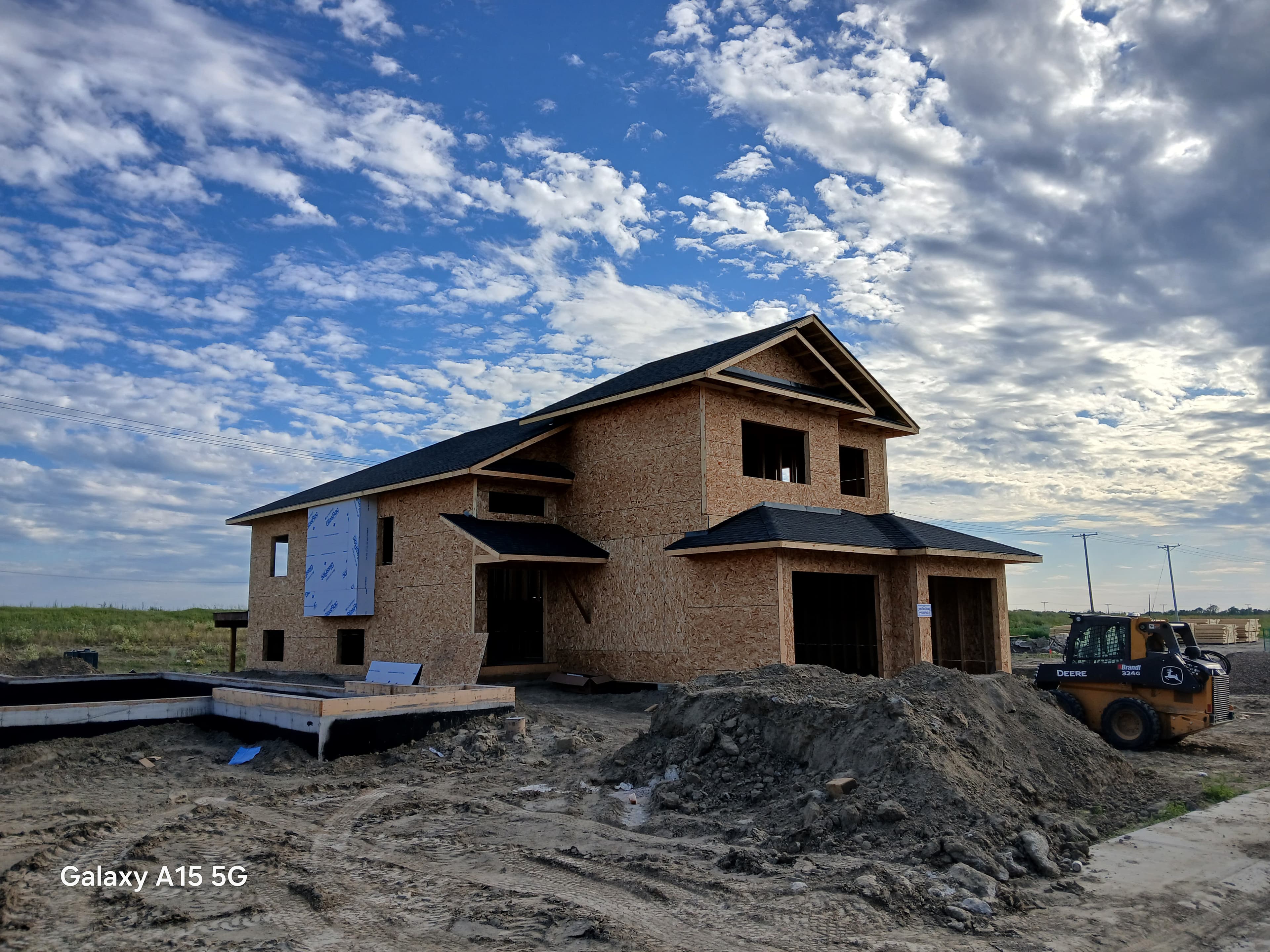 Two-story house under construction with OSB sheathing and a yellow skid steer on site.