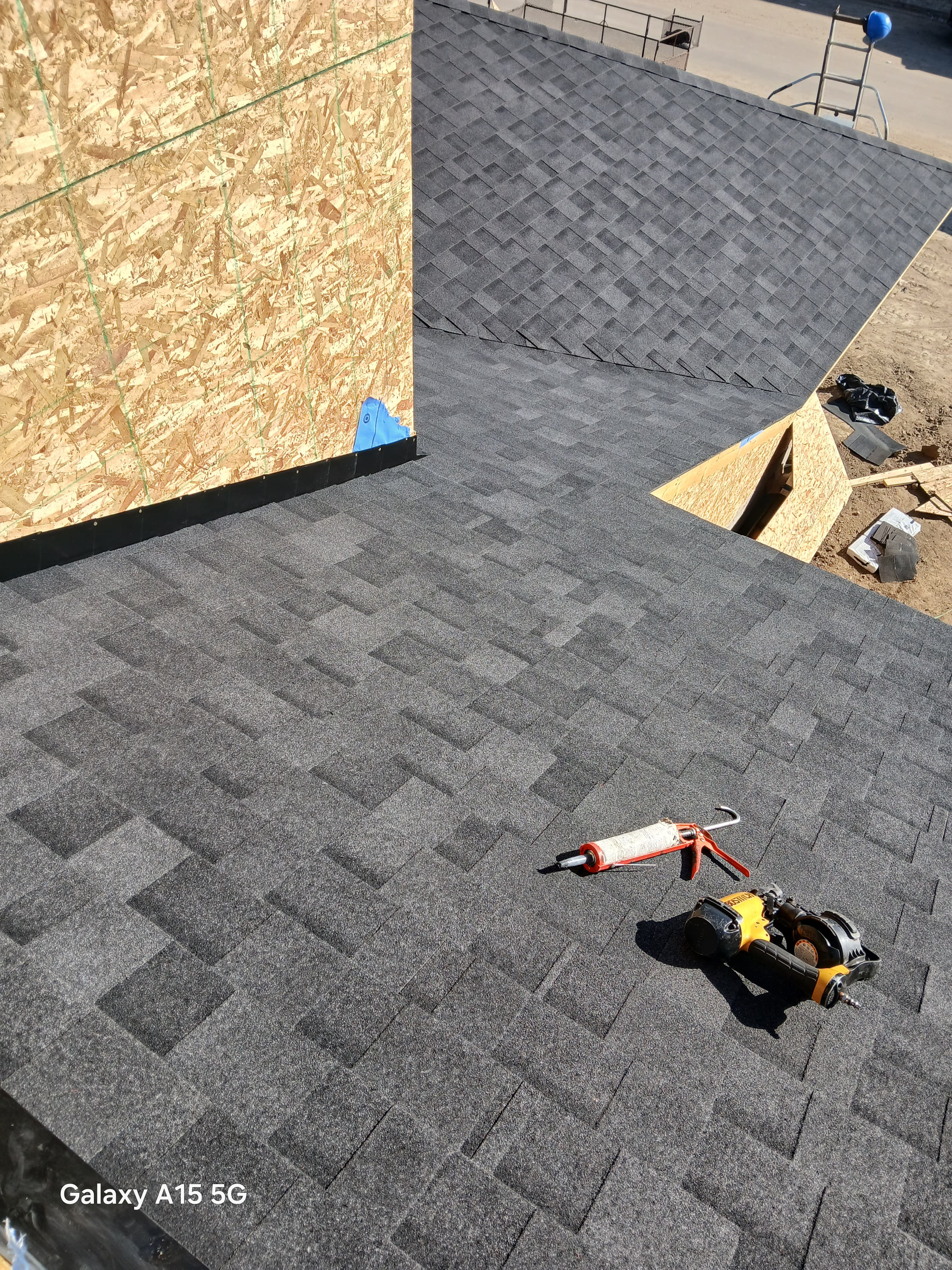 Yellow nail gun and orange caulking gun on dark gray shingles during roof construction.