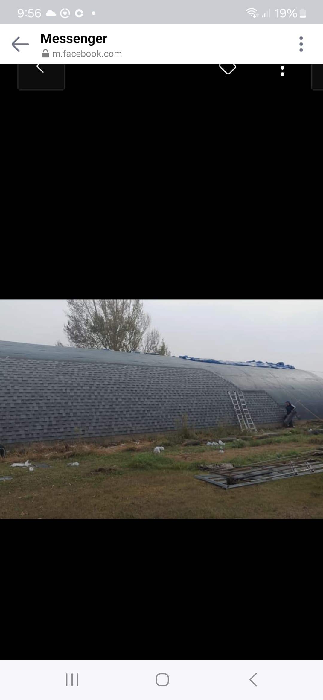 Worker installs grey asphalt shingles on a large curved building with a ladder nearby.