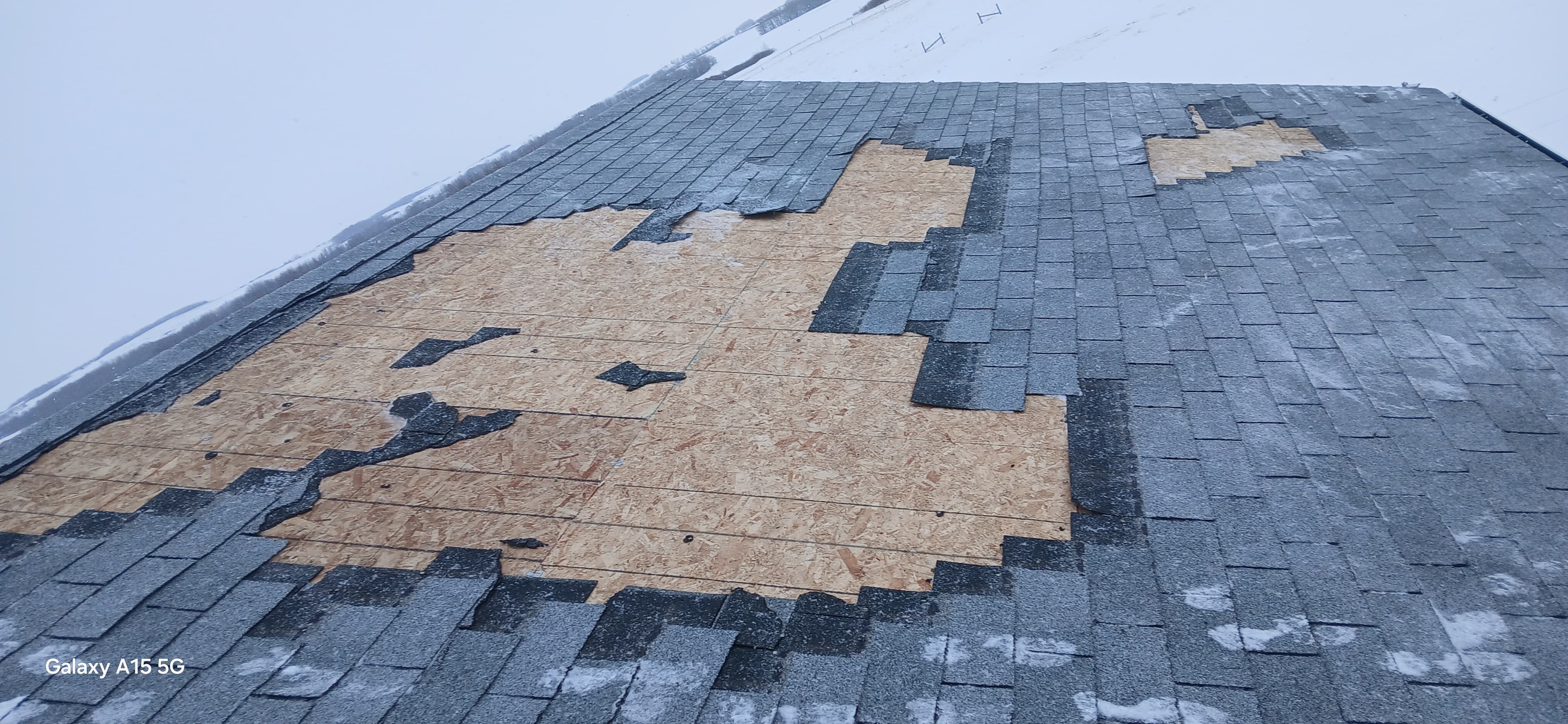 Damaged roof with missing grey shingles exposing wooden sheathing against a snowy winter background.