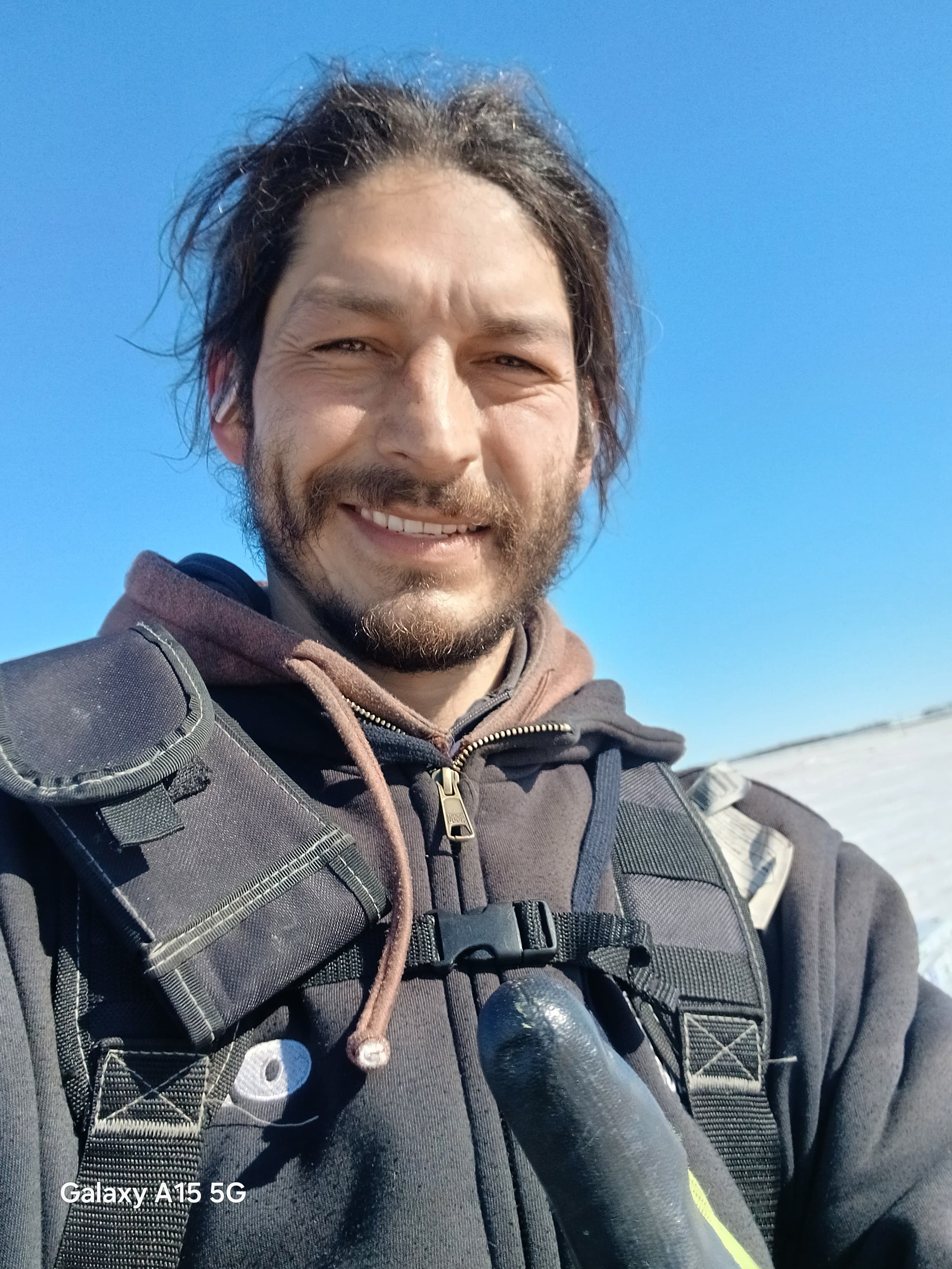 Bearded man smiling, wearing a black hoodie and tactical vest against a clear blue sky.