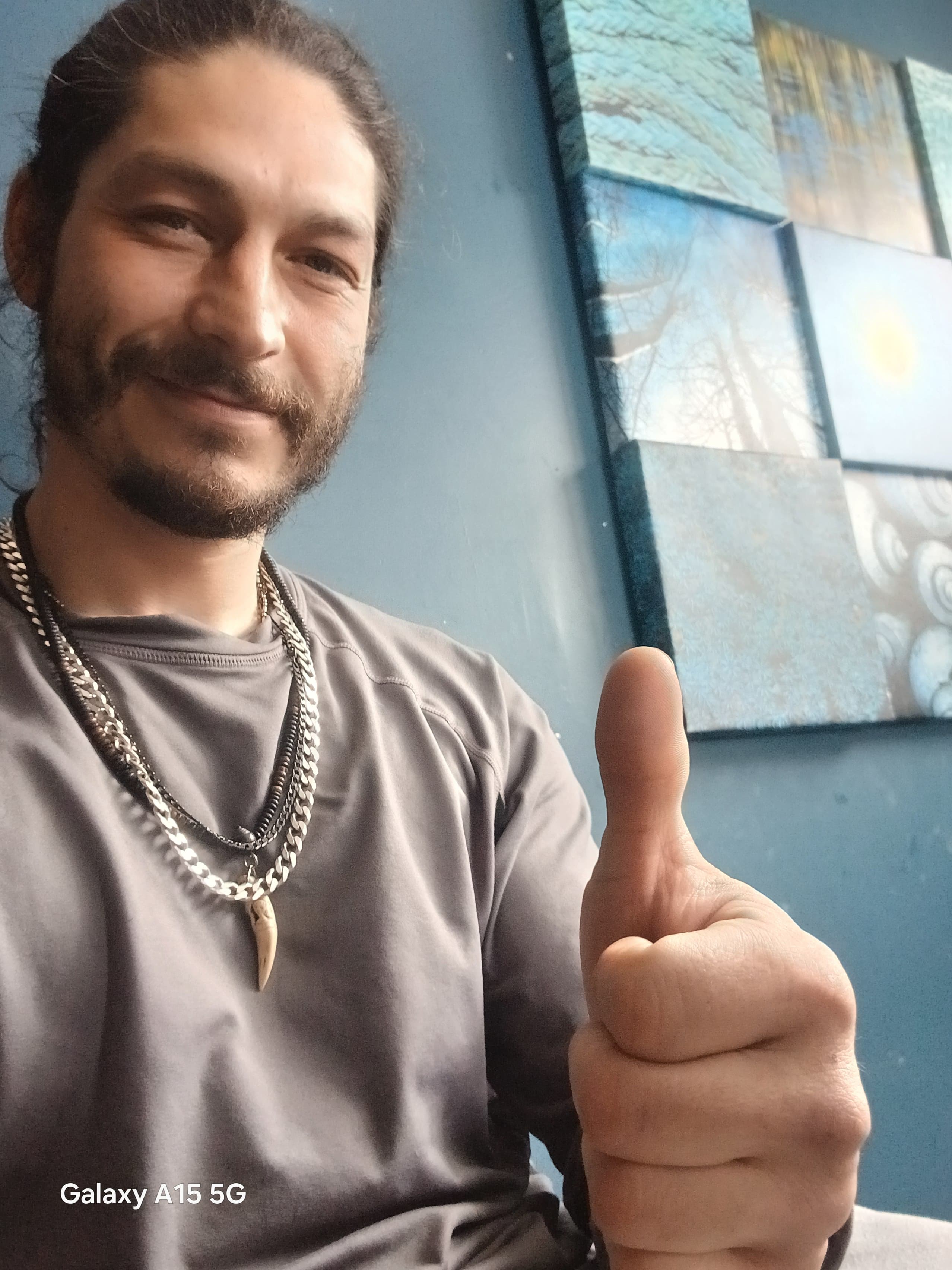 A bearded man gives a thumbs up, wearing silver necklaces and a grey shirt.