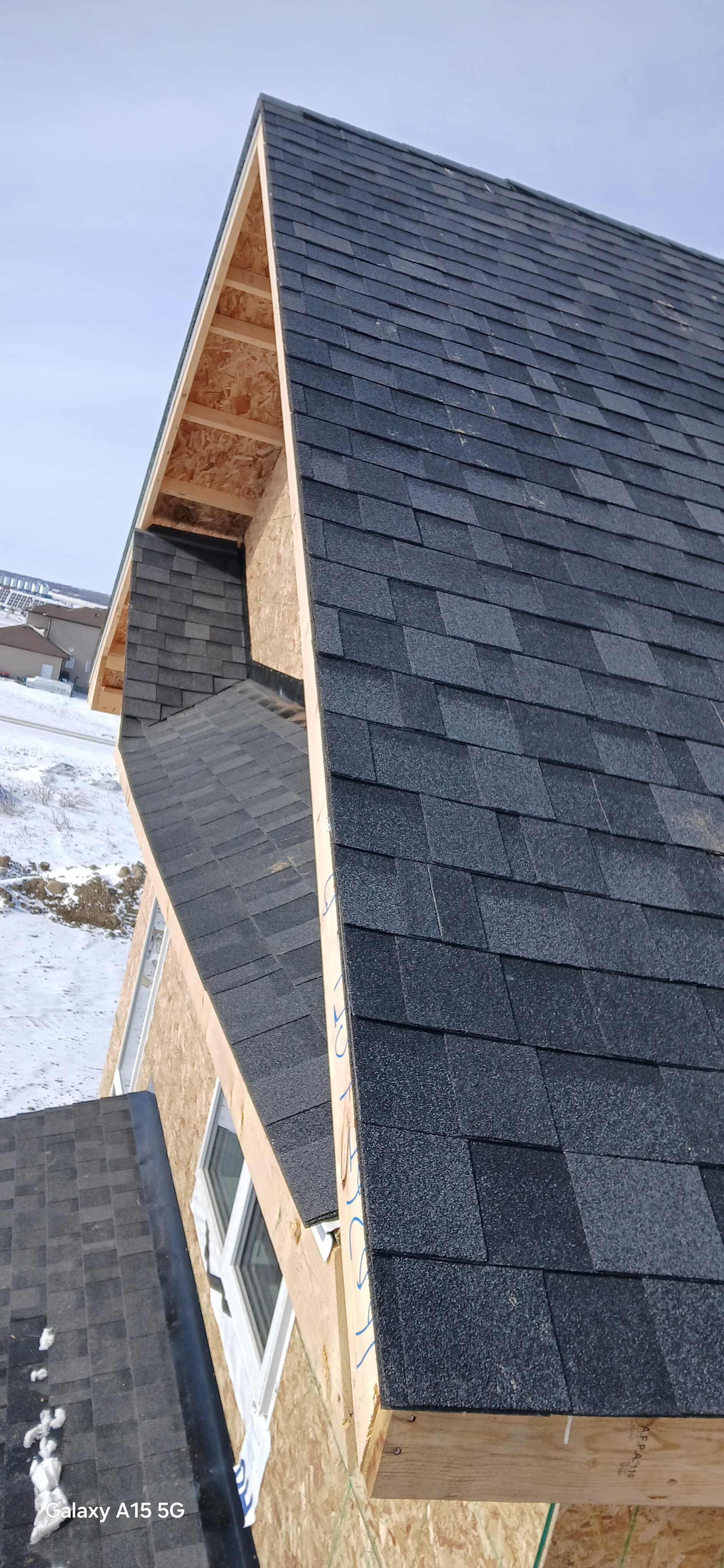 Dark grey asphalt shingles on a wooden roof frame under construction with snow visible below.