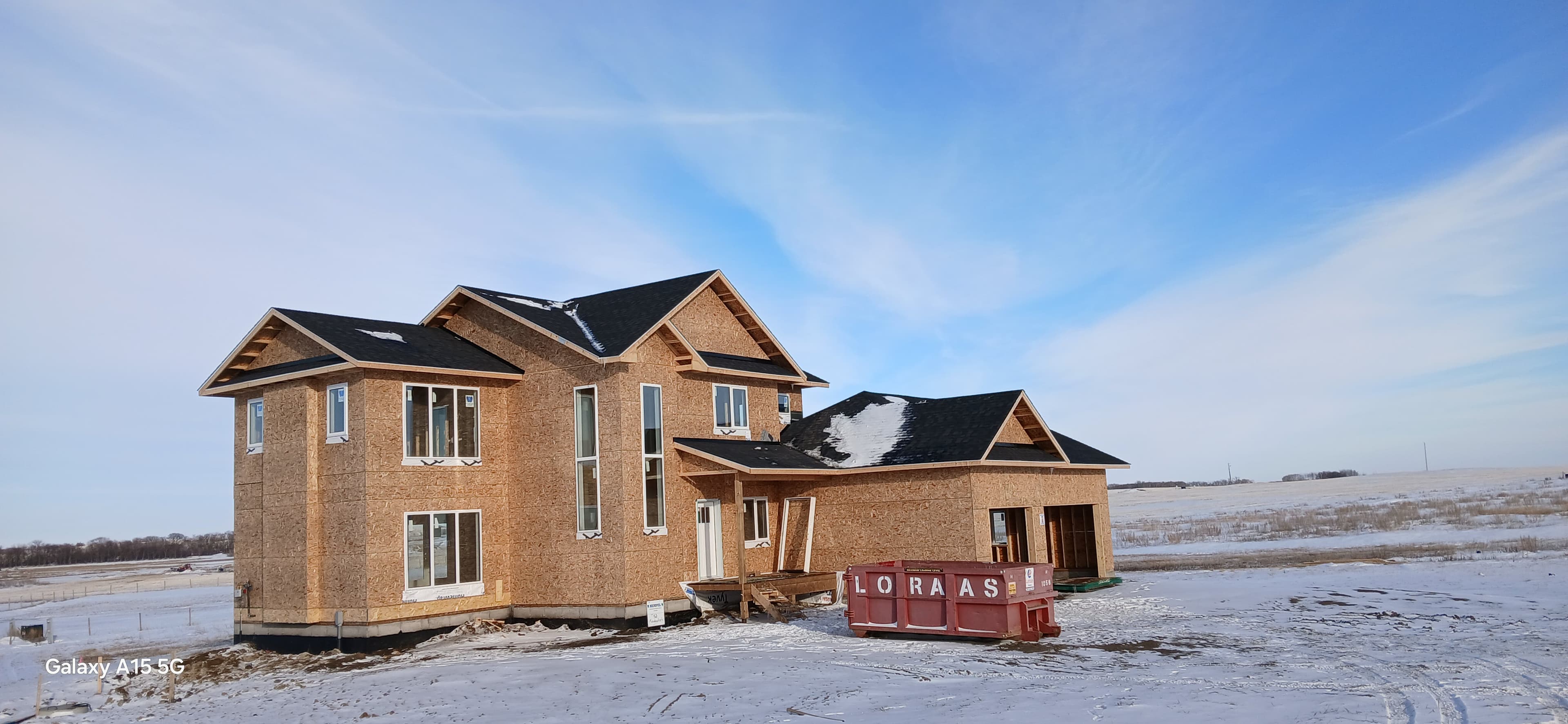 Unfinished house with plywood sheathing and black roof in a snowy, flat winter landscape.
