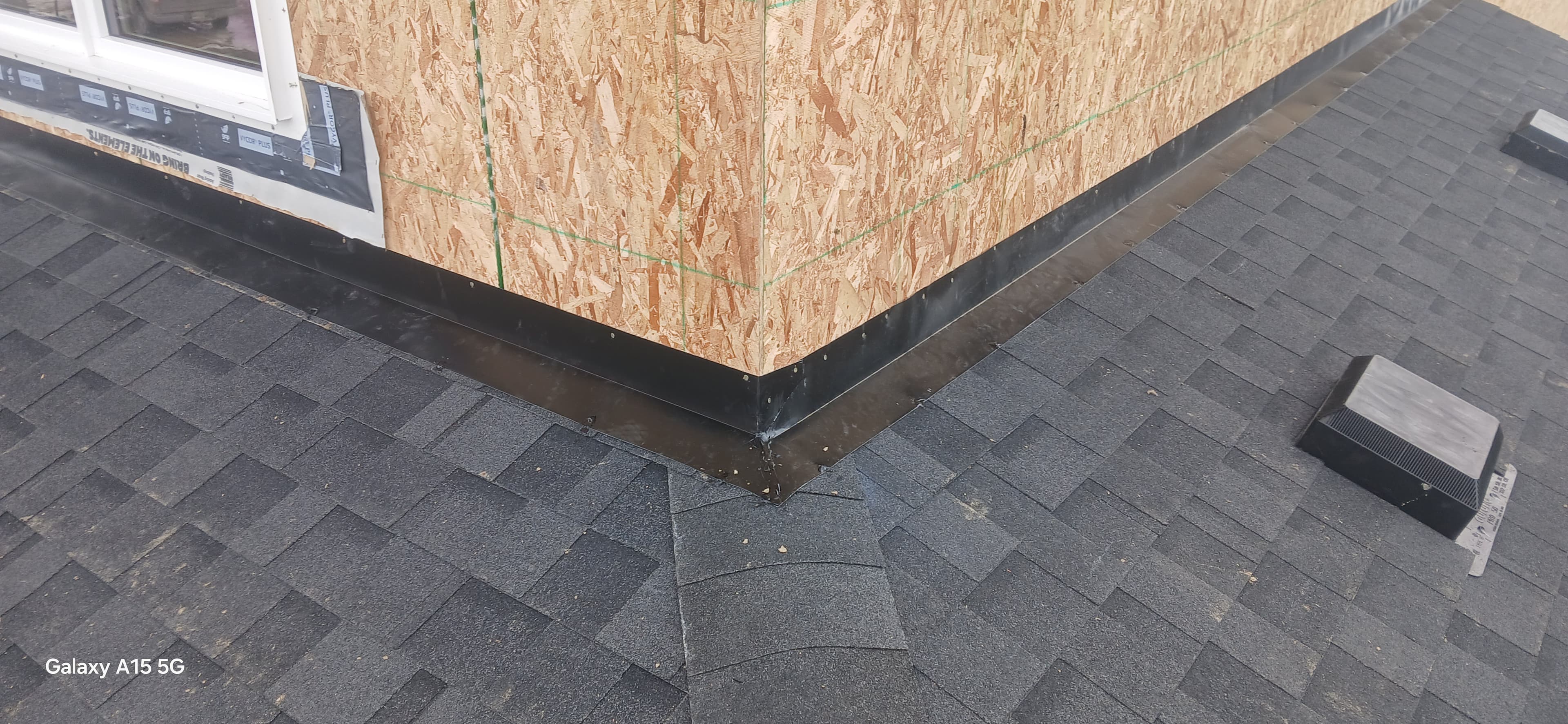 Black metal flashing at the intersection of dark grey asphalt shingles and an OSB wall.