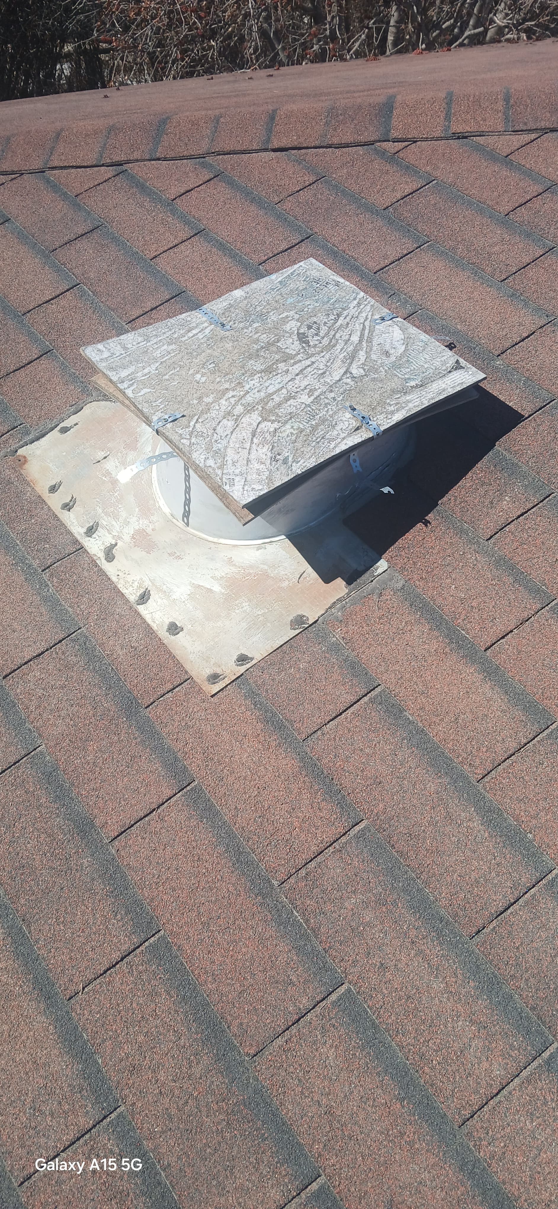 Weathered square cover partially obscuring a circular vent on a brown asphalt shingle roof.